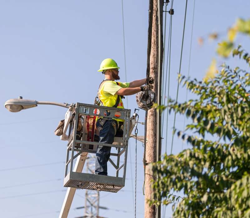 Worker in a hard hat and high-visibility shirt stands in a bucket lift, handling equipment on a utility pole against blue sky