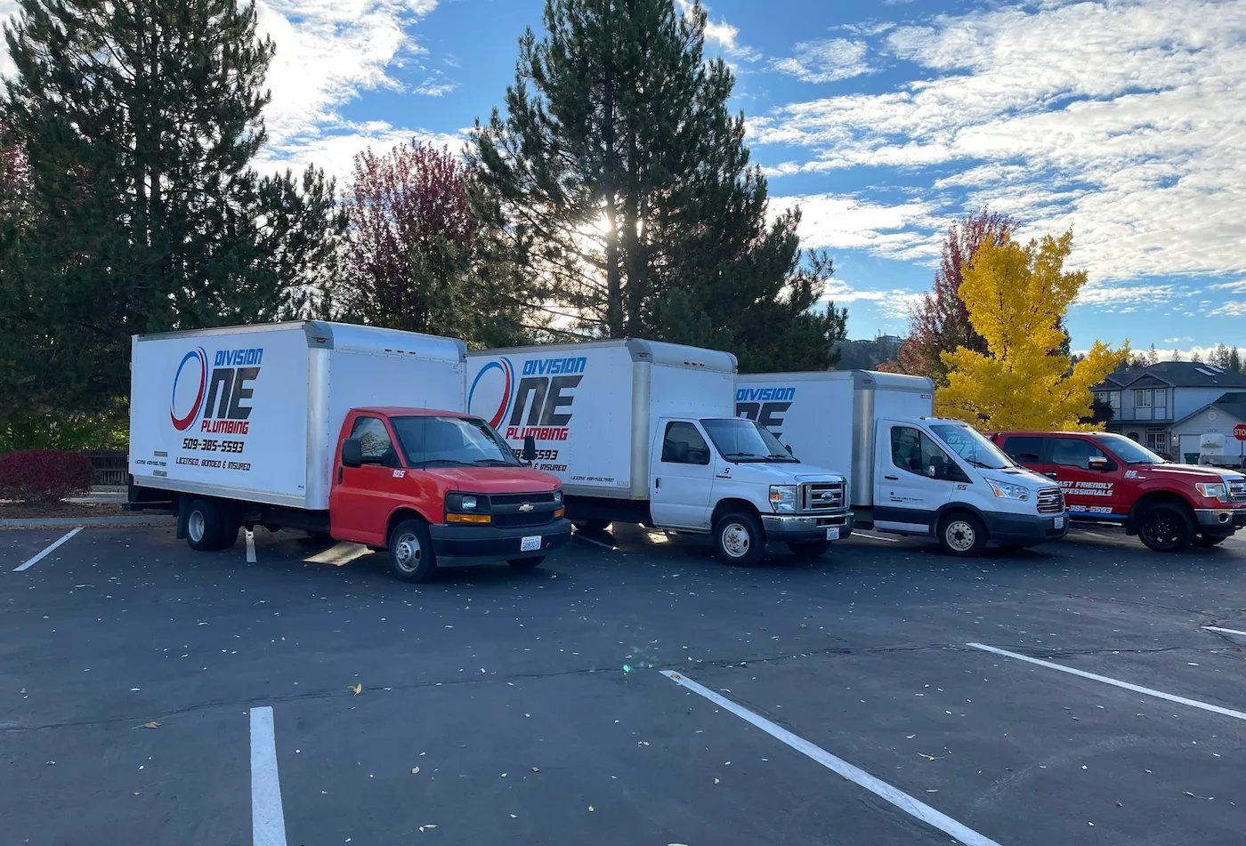 Four Division One Plumbing box trucks parked in a row in a lot, lit by low sun and fall trees