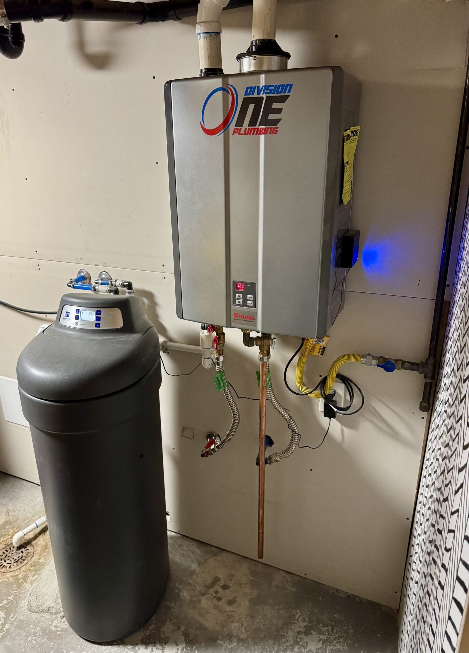 Wall-mounted tankless water heater with Division One Plumbing logo installed beside a water softener in a utility room