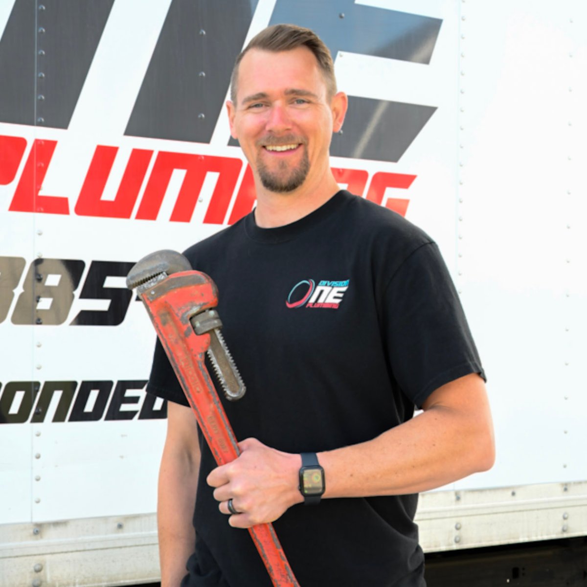 Smiling plumber in a Division One Plumbing shirt holds a large red pipe wrench in front of a company truck