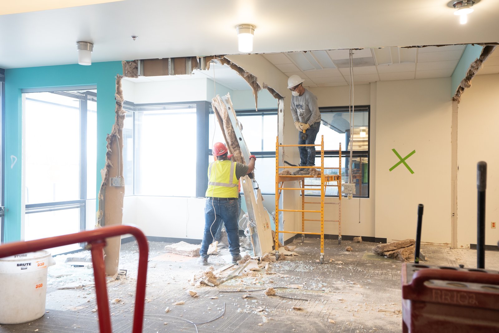 Workers open a wall and ceiling in a teal-painted office, with a scaffold and debris on the floor