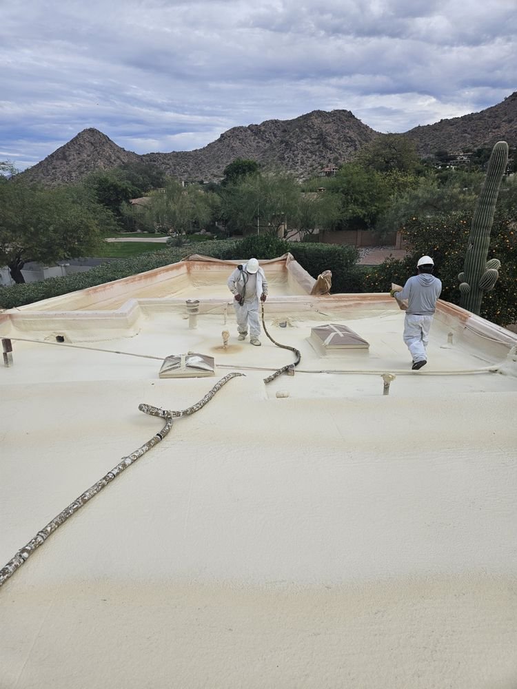 Two applicators in white protective suits and hard hats spray foam across a flat roof with desert mountains behind