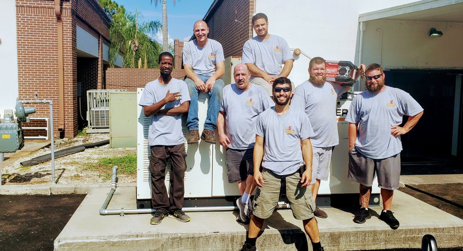 Seven Scruggs Electric crew members in matching gray T-shirts posing on and around a large outdoor generator next to a brick building