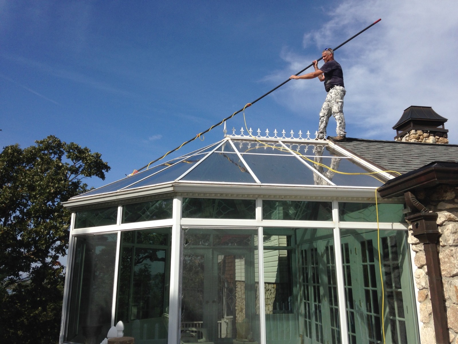 A man stands on a shingled roof and reaches across a glass conservatory with a long cleaning pole