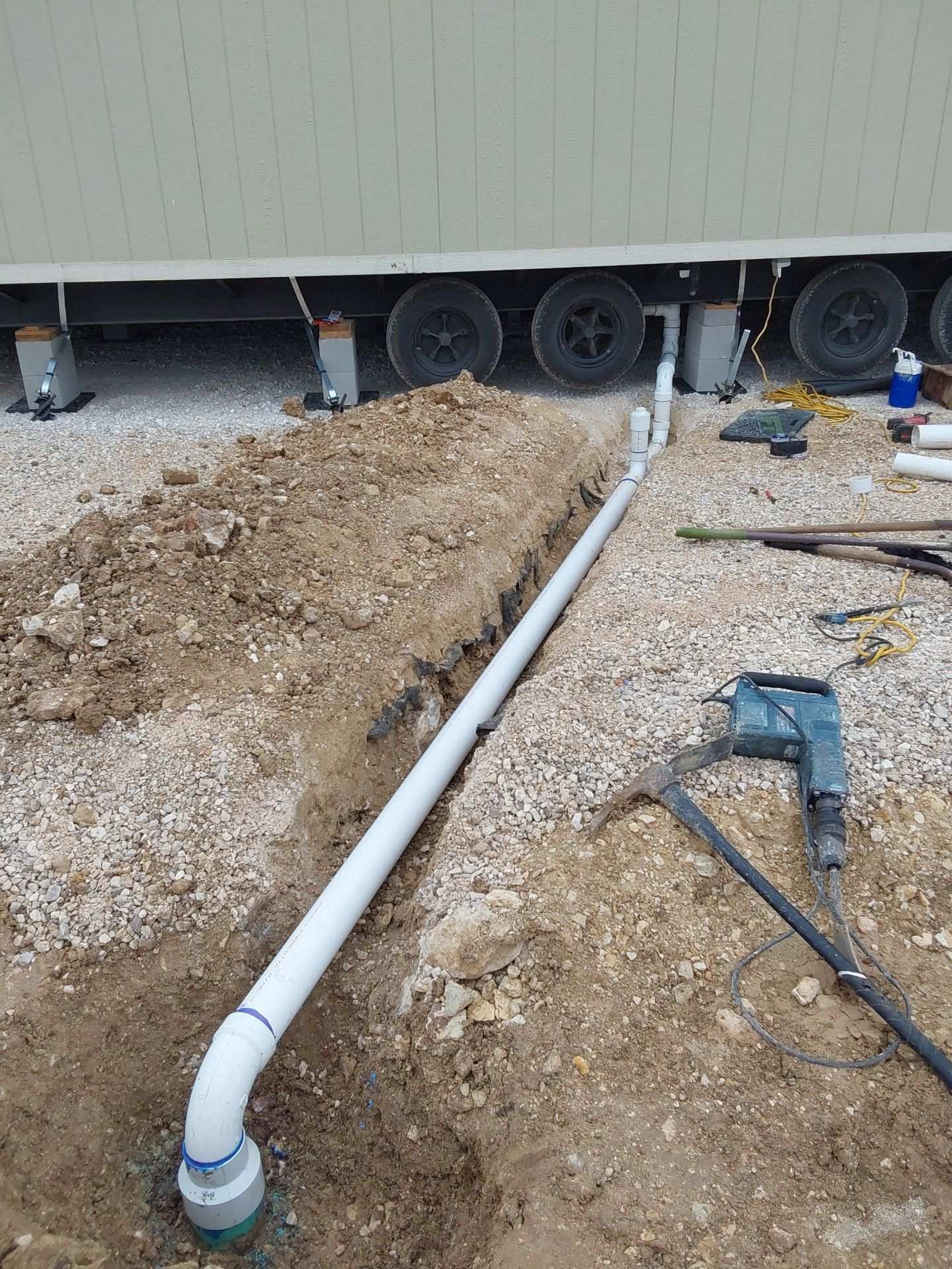 White PVC drain line laid in an open trench beside a trailer, with a jackhammer resting on the gravel