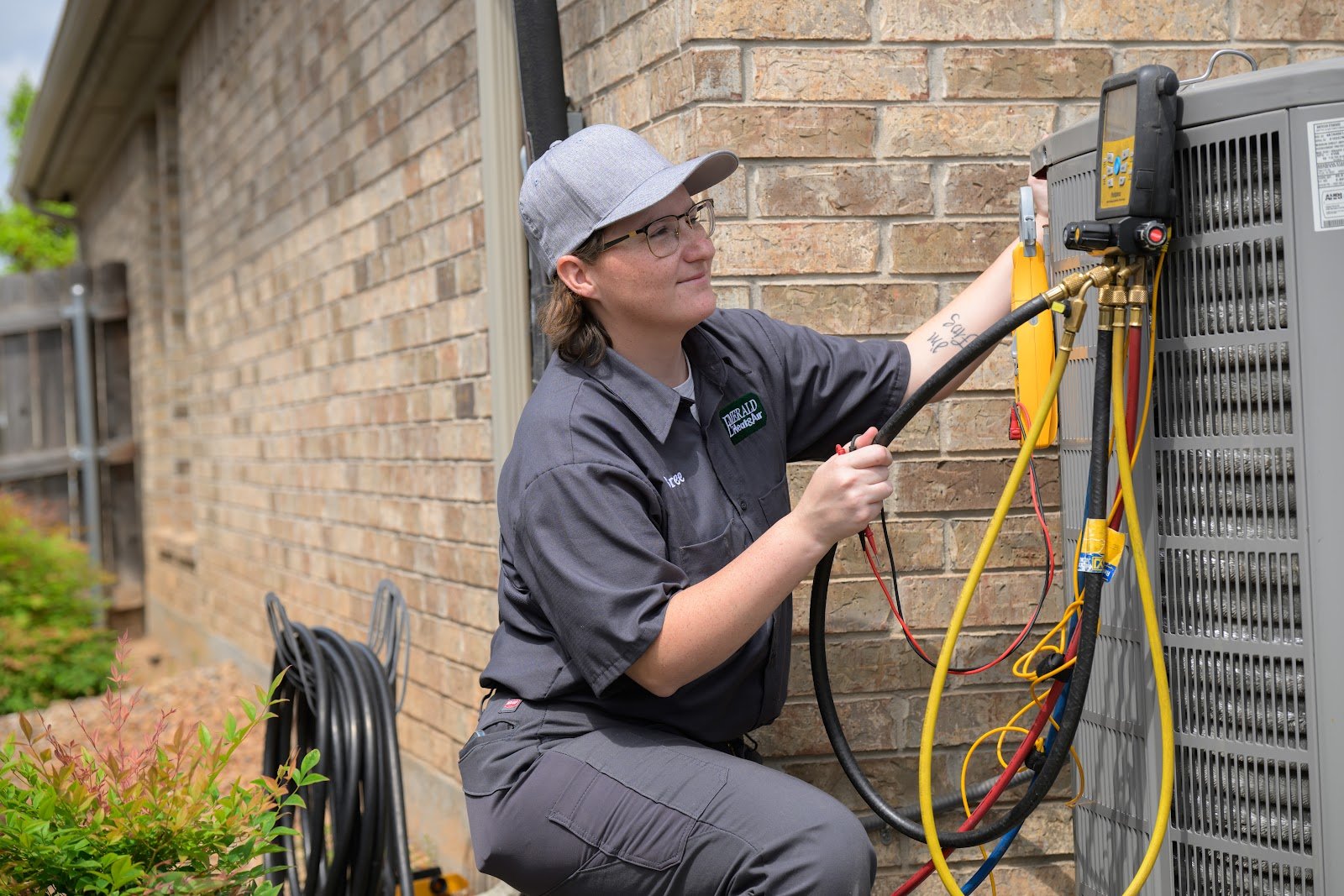 An Emerald technician in a gray work shirt and cap crouches beside an outdoor air conditioning unit with refrigerant gauges and hoses attached
