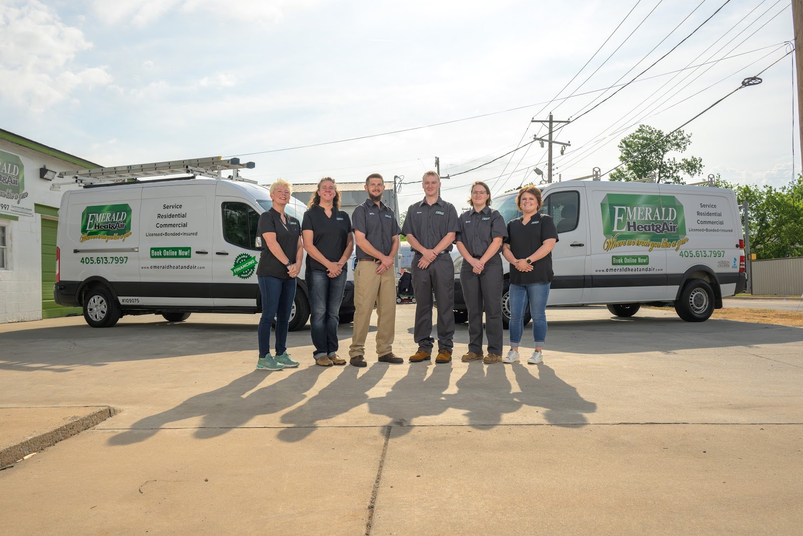 Six Emerald Heat & Air employees in company shirts stand in a parking lot between two white service vans with the green Emerald logo