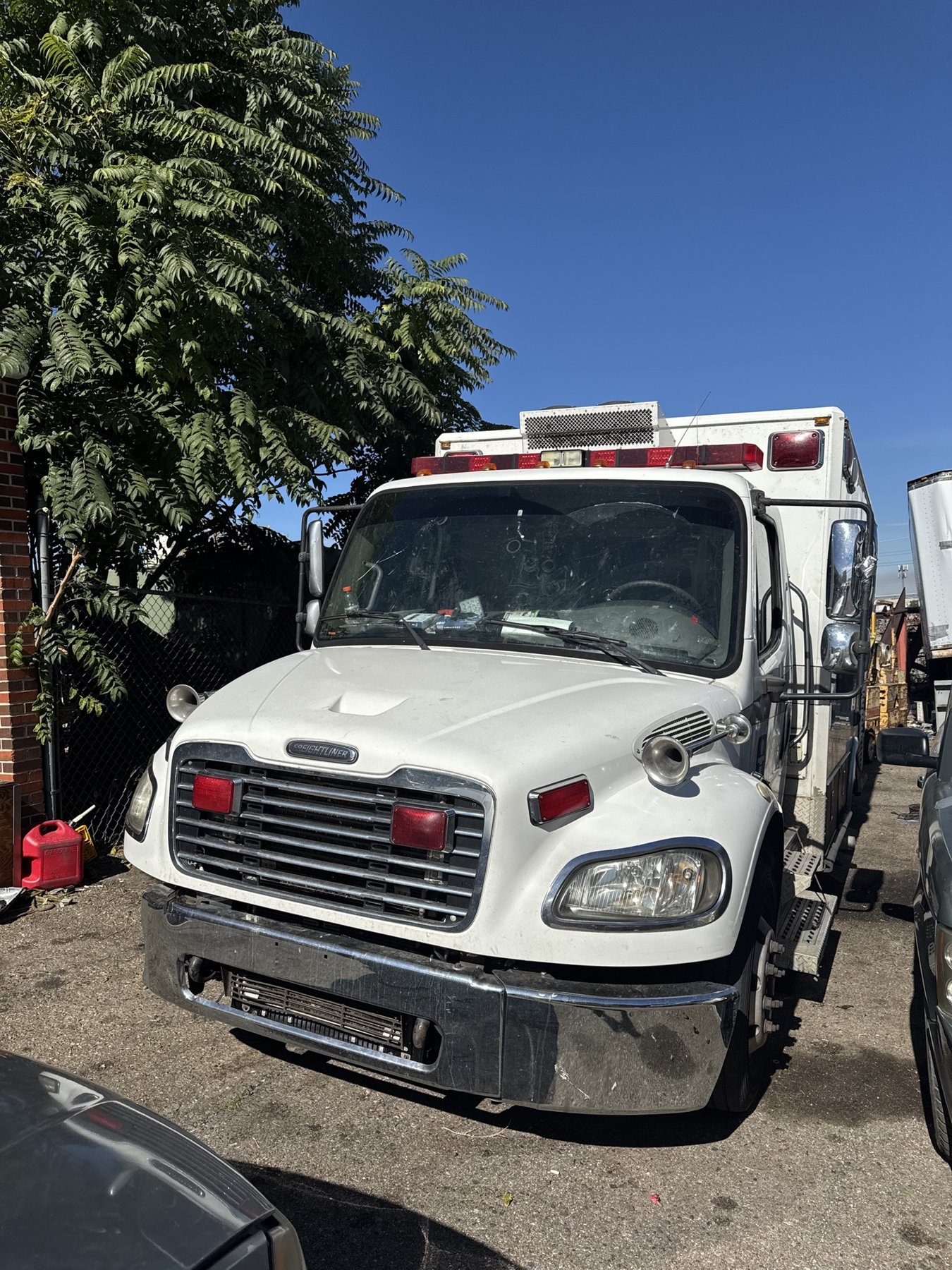 A white Freightliner ambulance converted into a refrigeration service truck, parked beside a tree