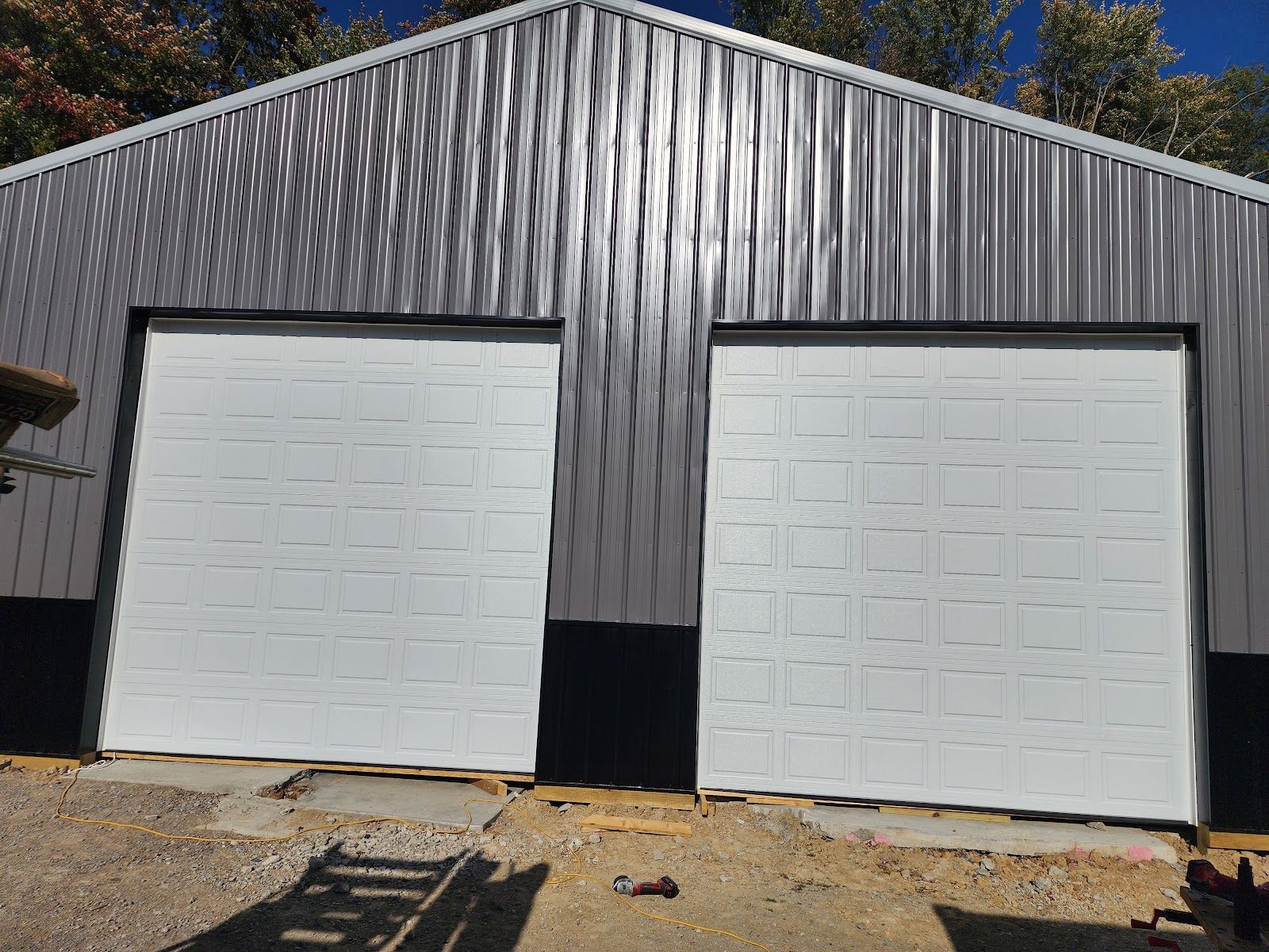 Two new white sectional garage doors set in the gray steel wall of a newly built pole building, with an extension cord, tools and scrap wood on the gravel in front