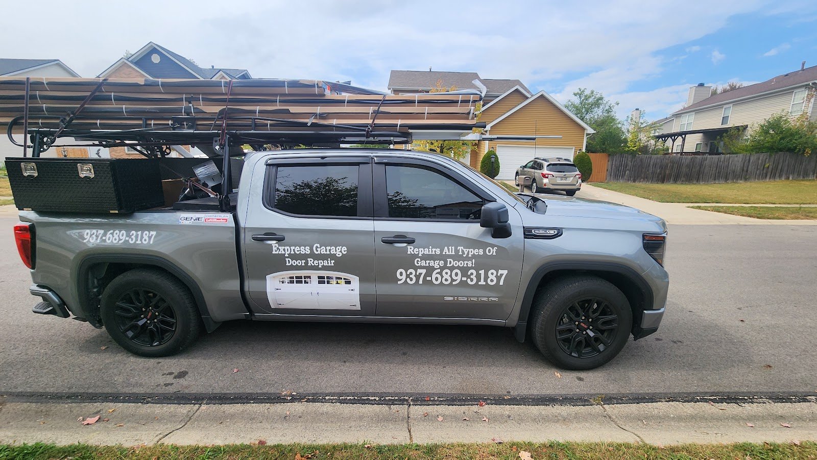 A gray GMC pickup lettered Express Garage Door Repair, with wrapped garage door sections strapped to a ladder rack over the bed, parked at a curb on a residential street