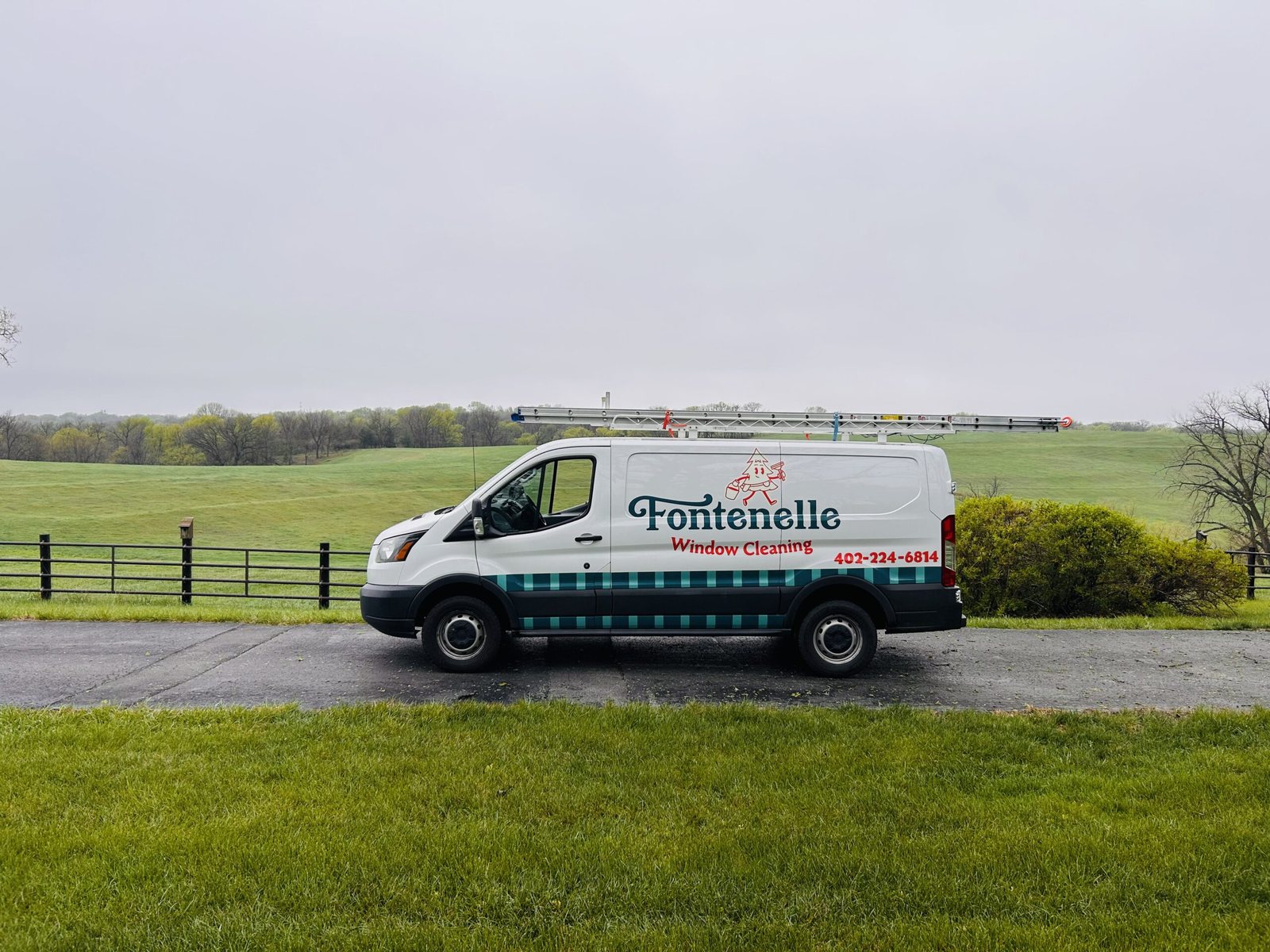A white Fontenelle Window Cleaning van with ladders on its roof rack, parked on a paved drive beside a green pasture