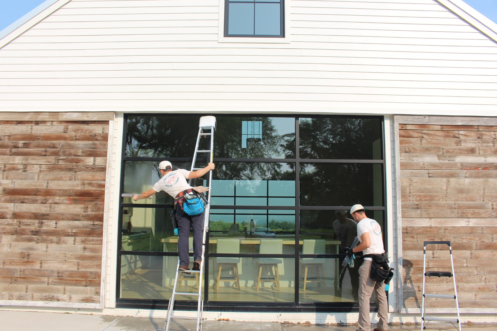 Two workers in white Fontenelle Window Cleaning shirts and caps clean a wall of large black-framed windows, one of them standing on a ladder