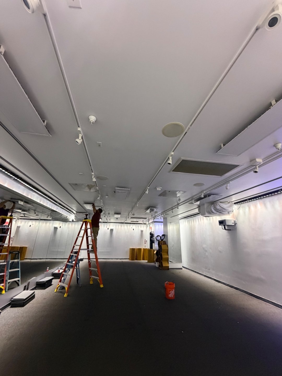 Electrician on an orange ladder working on the ceiling of a large empty commercial space with track lighting