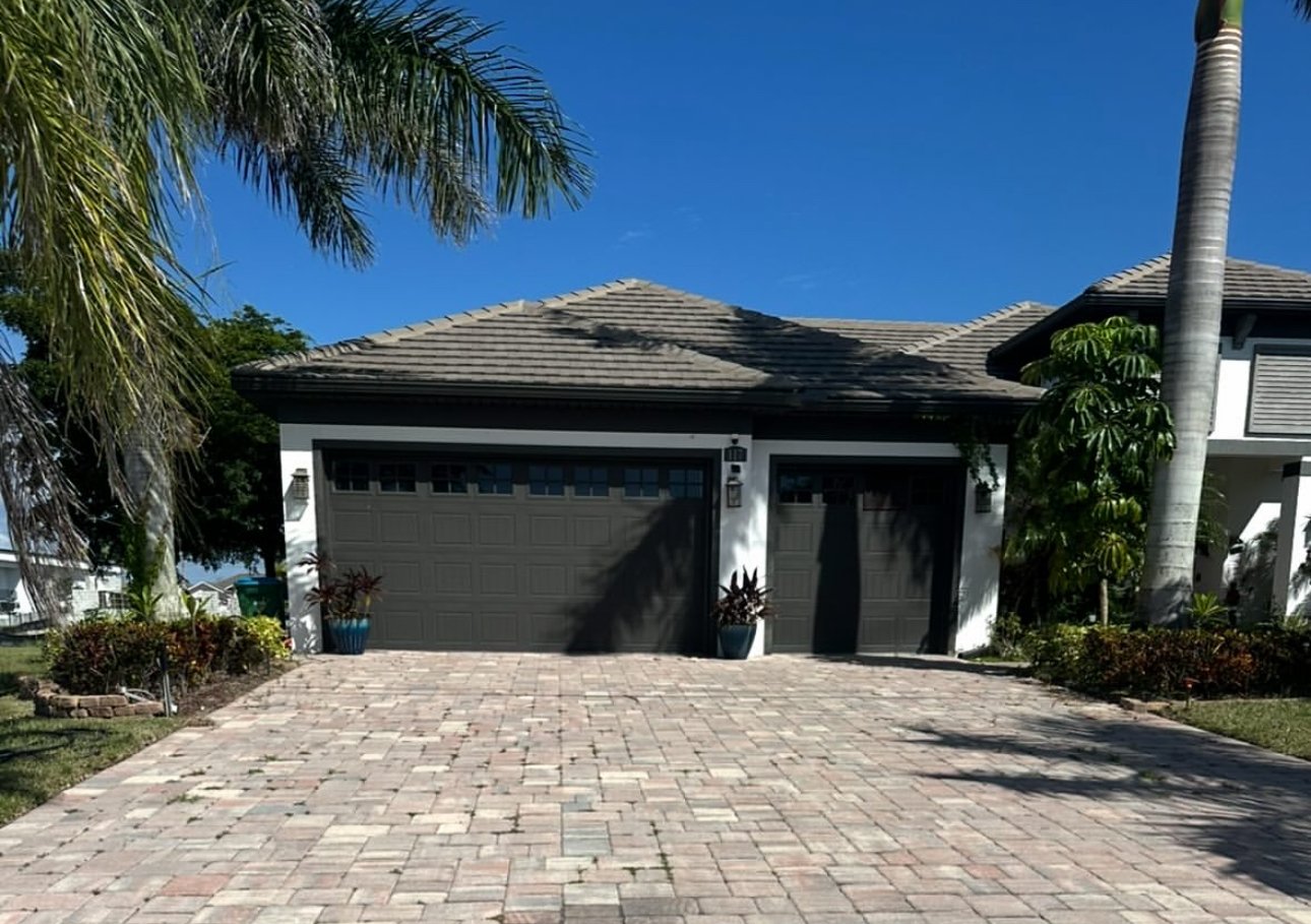 Two-car garage with dark gray paneled doors, a paver driveway and palm trees under a blue sky