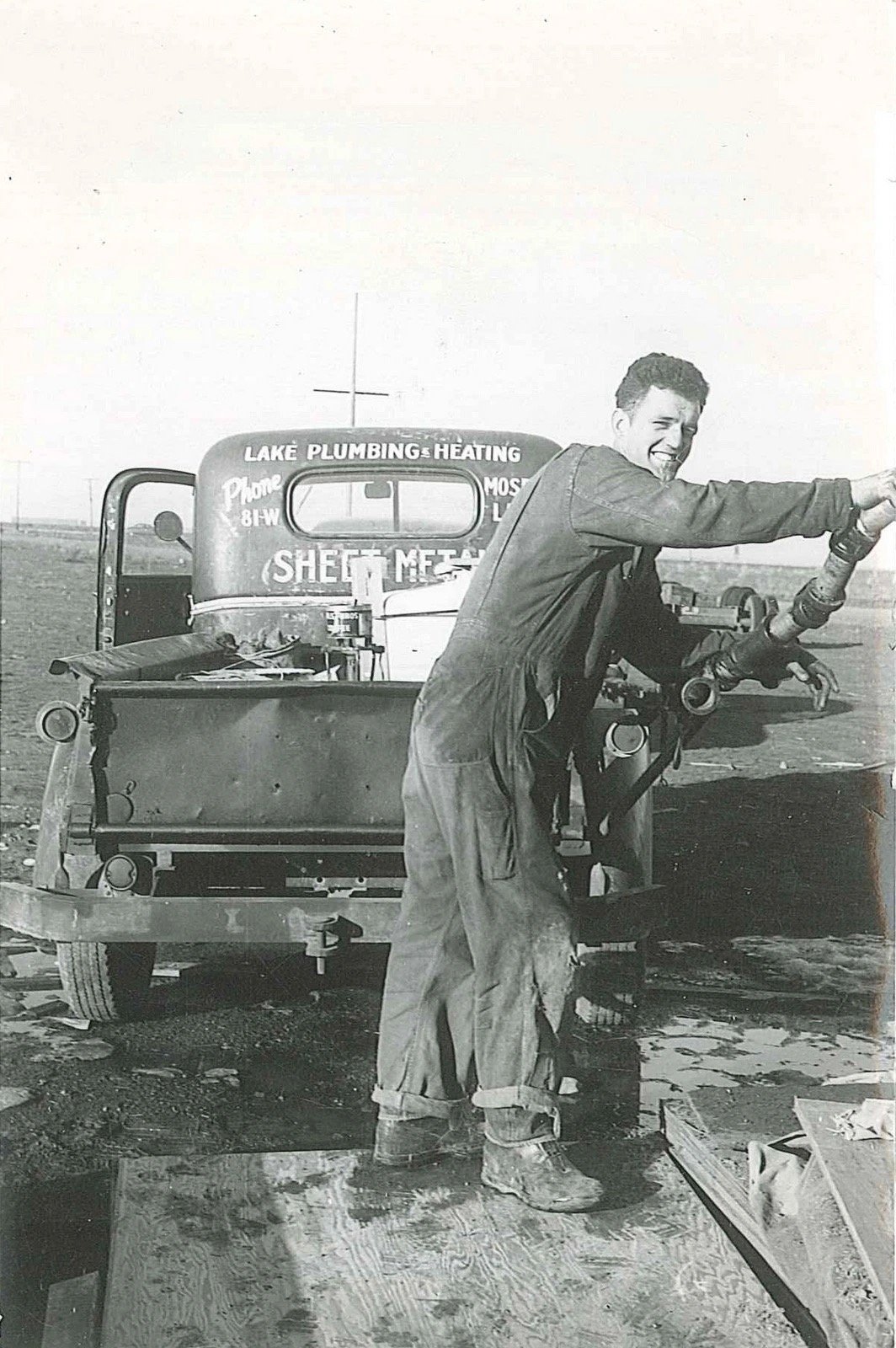 A black-and-white photo of a smiling young man in coveralls and work boots holding a length of pipe beside the back of an old truck lettered Lake Plumbing & Heating