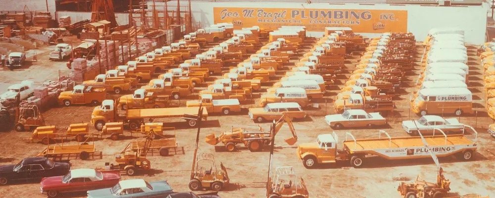 A faded color photo of a large yard filled with rows of yellow pickup trucks, vans, flatbed trailers and backhoes, with a long yellow sign on a building reading Geo M Brazil Plumbing Inc. Mechanical Contractors