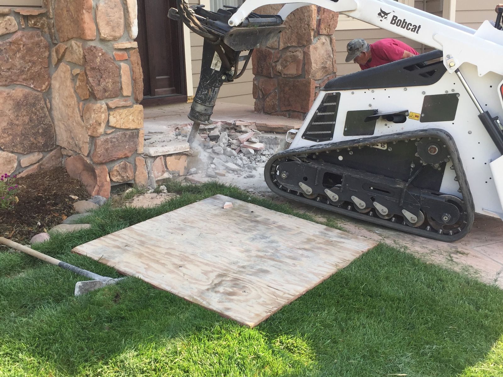 A small tracked loader with a hydraulic breaker breaks out an old step beside a stone-faced porch, a sheet of plywood laid on the lawn