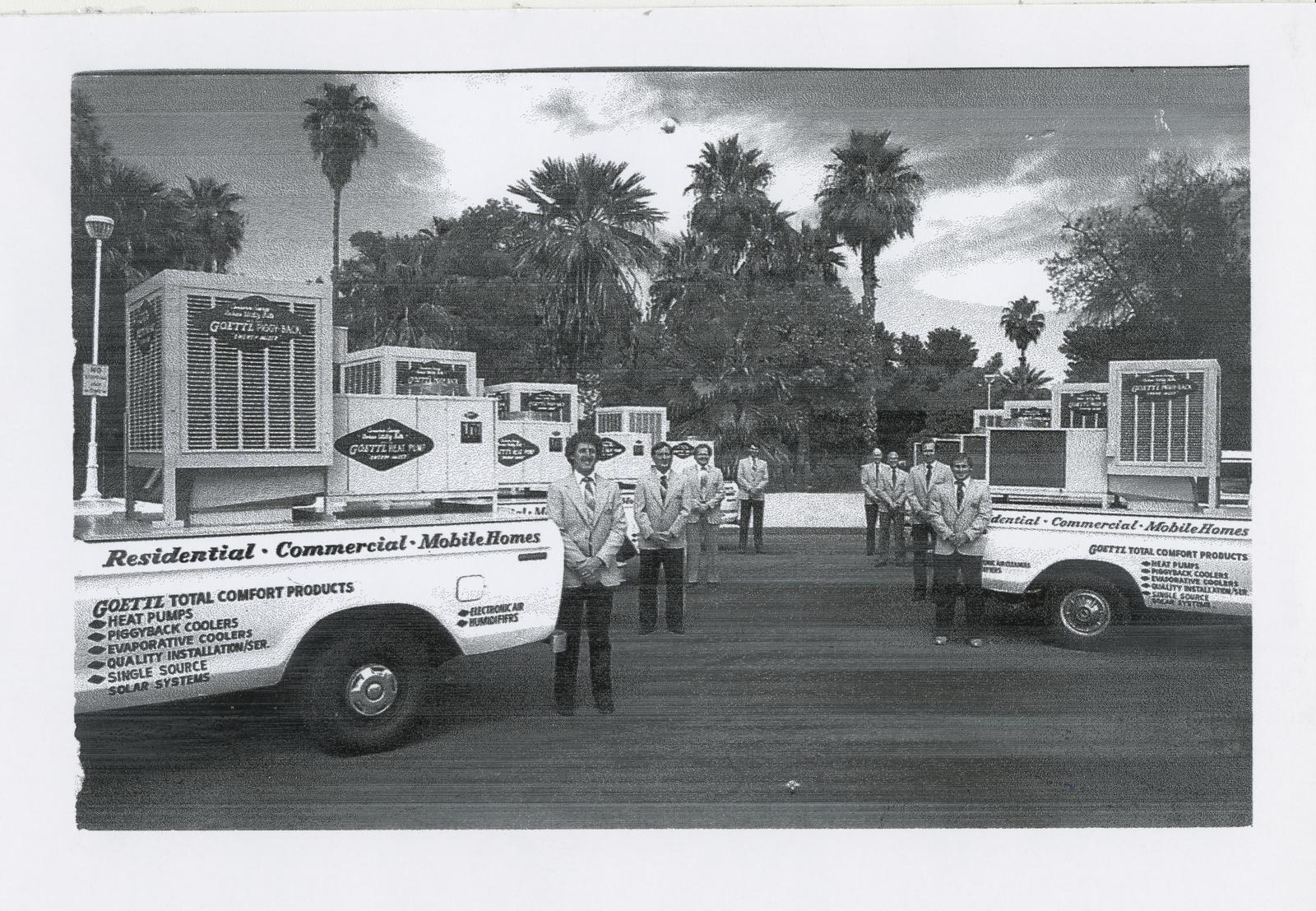 A black-and-white photo of men in suits standing beside two white Goettl pickup trucks loaded with evaporative coolers, with palm trees behind them