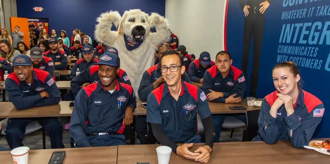 Goettl technicians in navy and red uniforms sit at long tables in a training room, smiling, with a person in a white dog mascot costume standing behind them