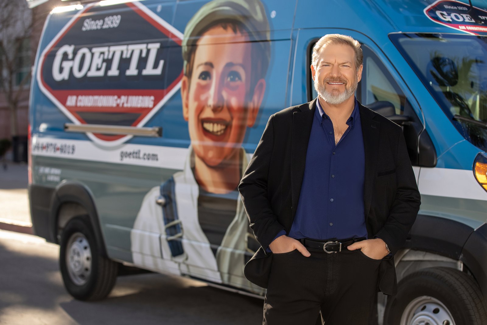 Ken Goodrich, a man with a gray beard in a dark jacket and blue shirt, stands in front of a blue Goettl Air Conditioning & Plumbing van painted with a smiling boy in a cap