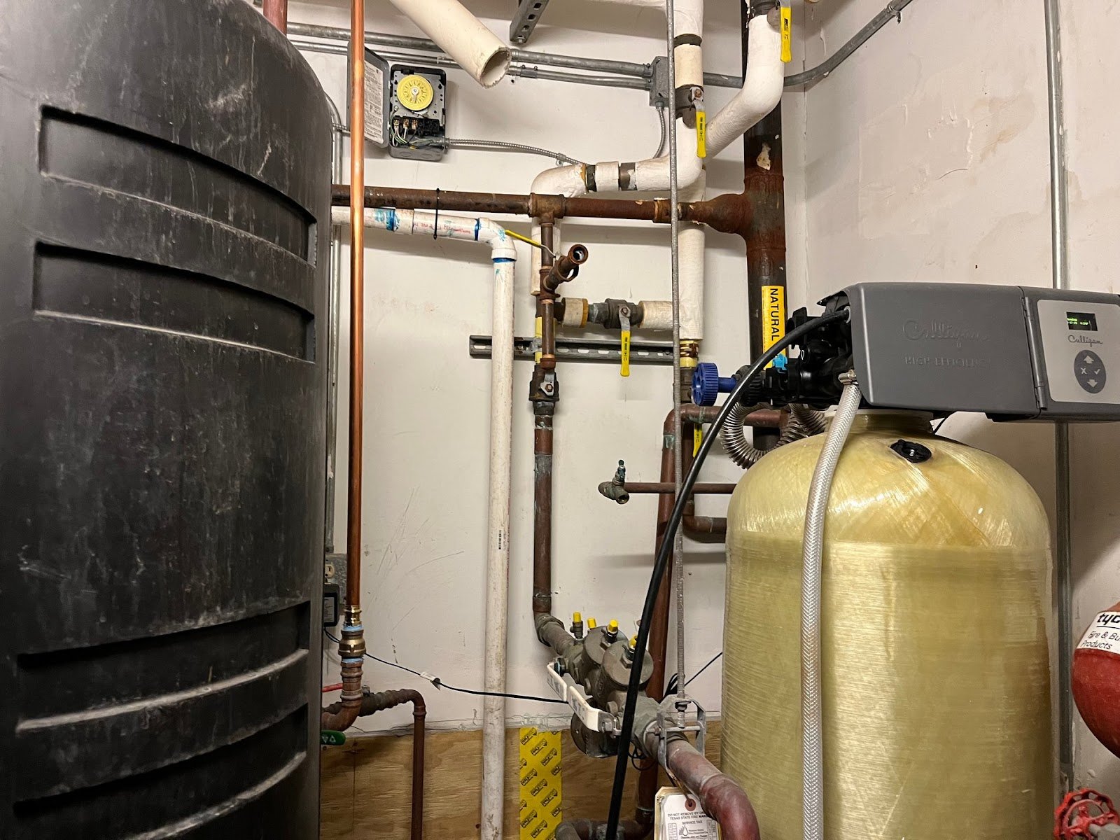 Commercial mechanical room with copper and PVC water piping, a black storage tank and a yellow water softener tank