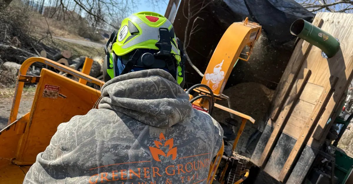 Crew member in a bright green helmet and Greener Grounds sweatshirt stands at an orange wood chipper