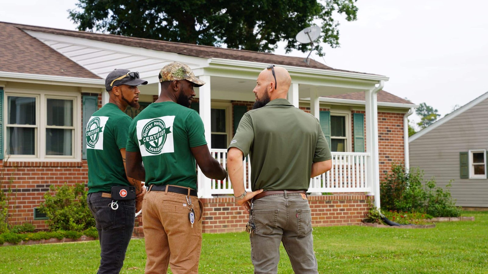 Three men in green certified shirts and work gear stand on a lawn talking in front of a brick house