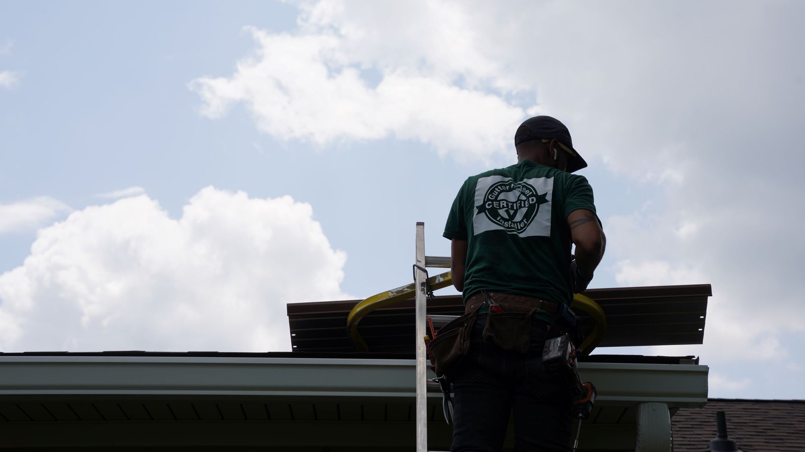Installer in a green certified crew shirt stands on a ladder at the roof edge, carrying a gutter cover section