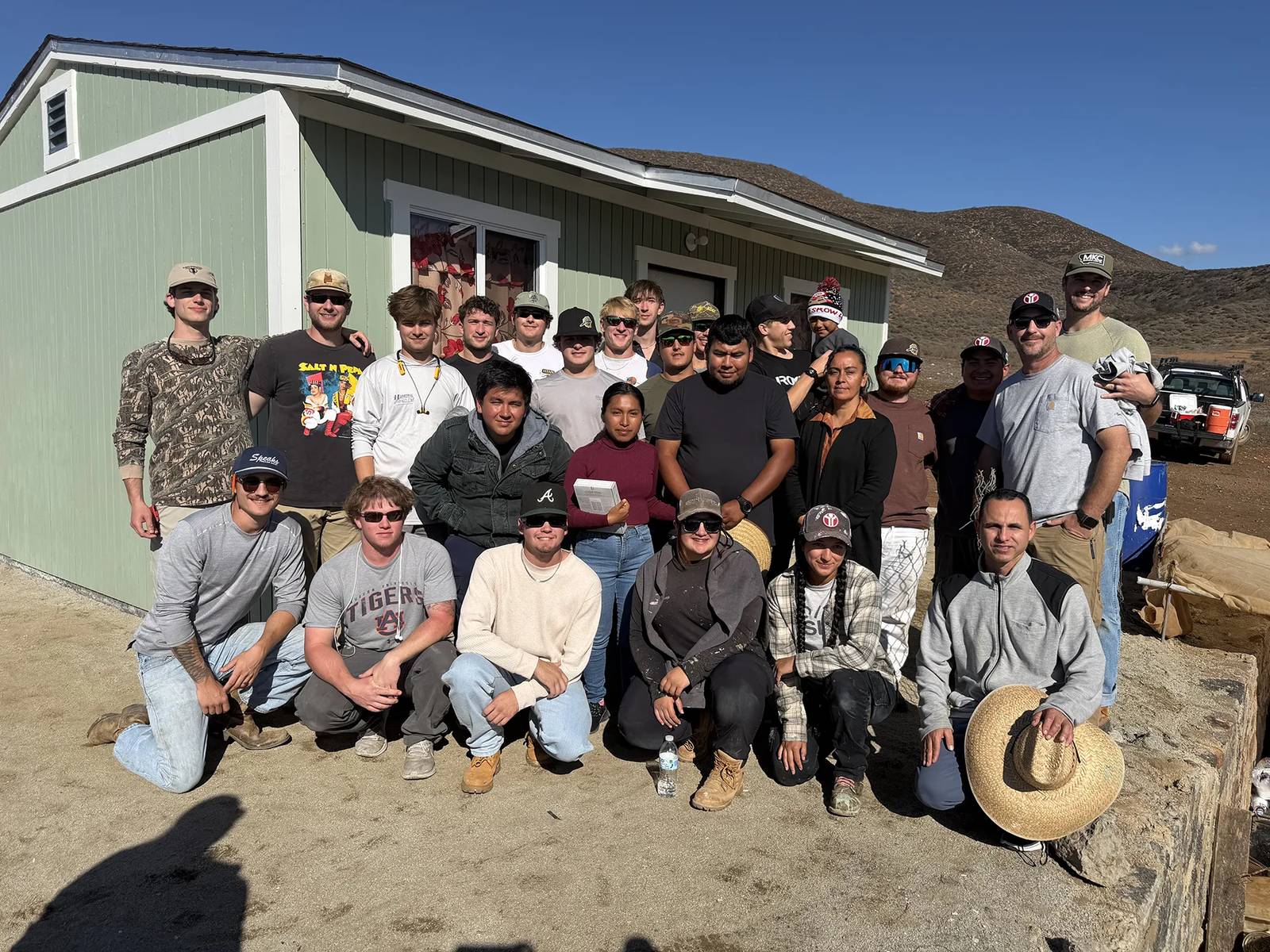 A group of about two dozen people pose in front of a newly built pale green house in dry, hilly country