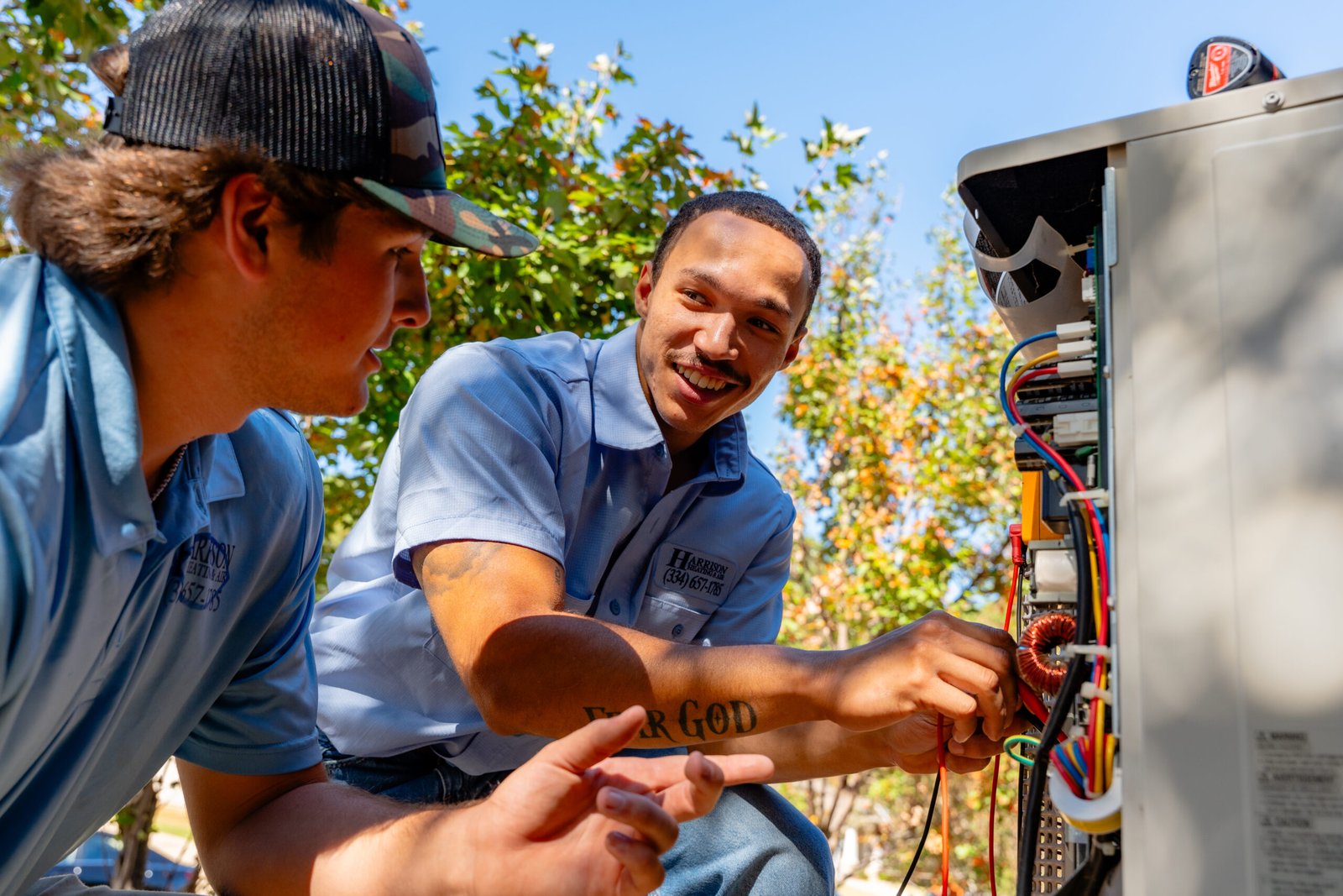 Two technicians in blue work shirts embroidered with the Harrison logo work on the wiring inside an outdoor air conditioning unit
