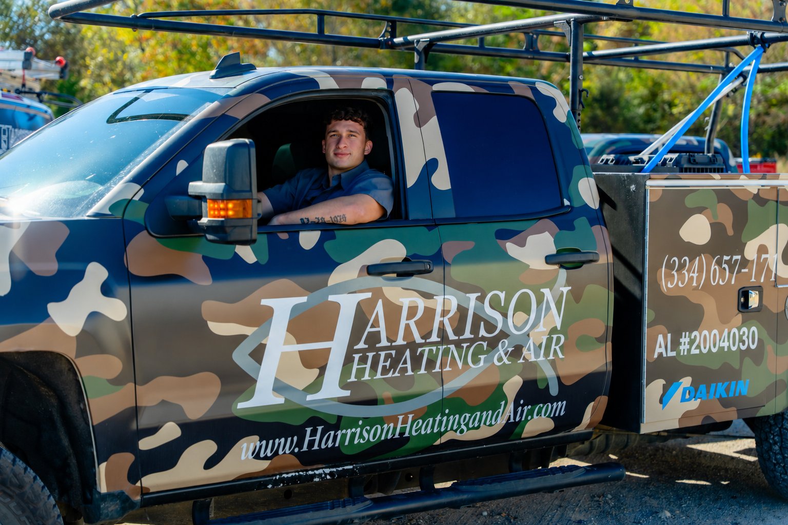A pickup in a camouflage wrap lettered Harrison Heating & Air, with a young man in a work shirt in the driver's seat