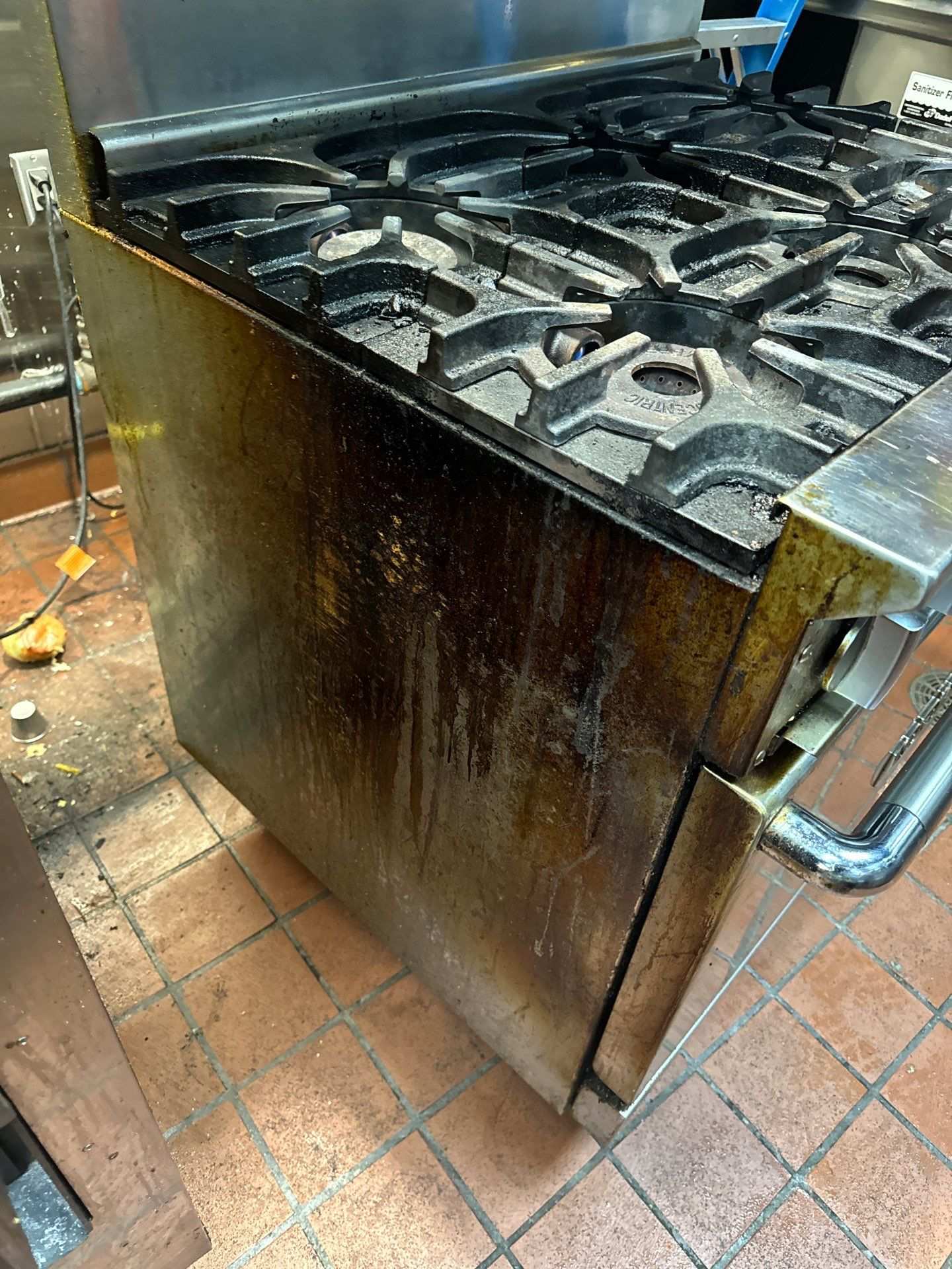 Grease-stained commercial gas range with cast iron burners on a tiled kitchen floor, awaiting deep cleaning