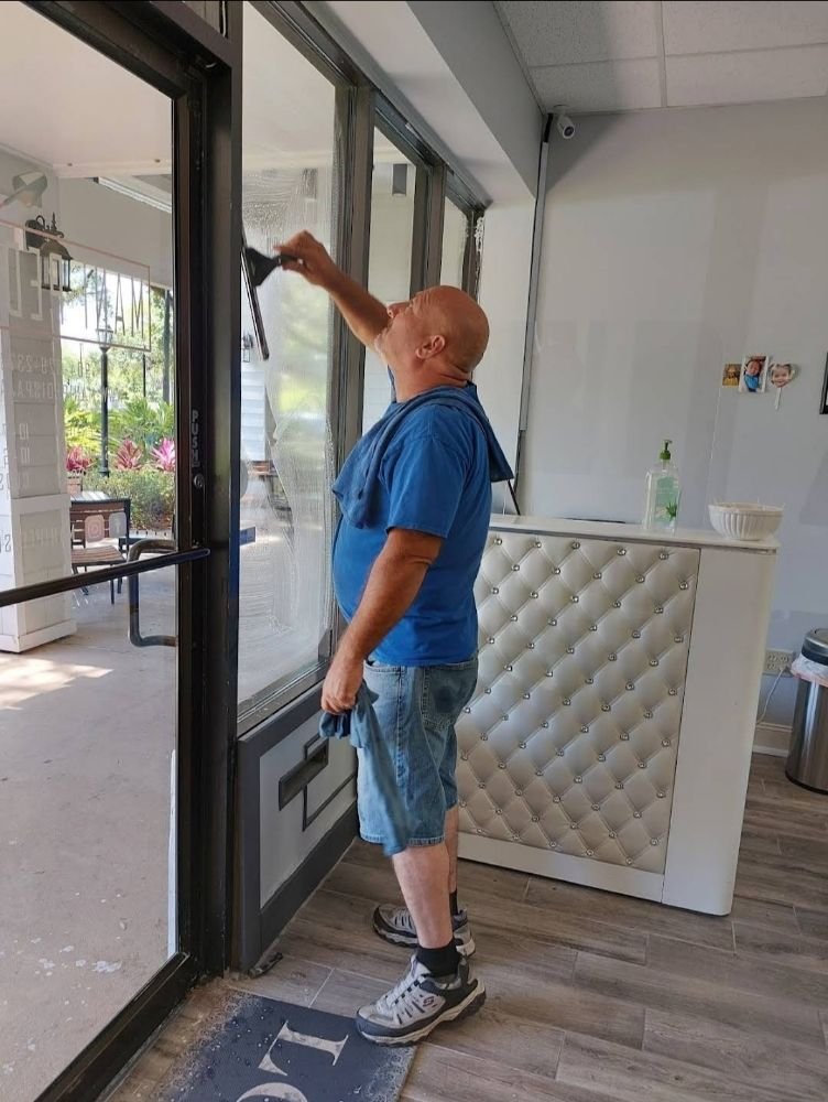 Window cleaner in a blue shirt and denim shorts squeegeeing a storefront glass door inside a small shop