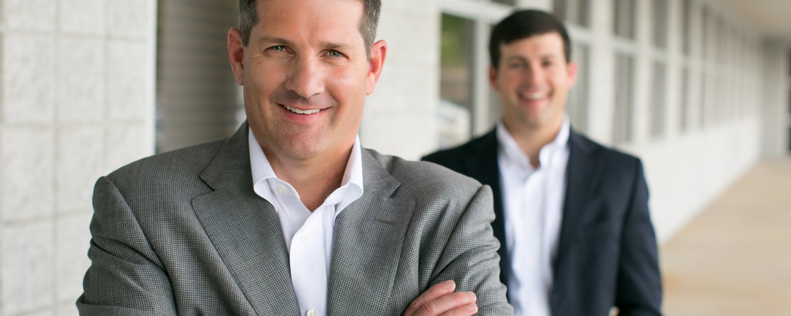 Jimmy Hiller in a gray jacket and white open-collar shirt, arms crossed and smiling, with his son Jimmy Hiller Jr. in a navy blazer standing behind him in a covered walkway