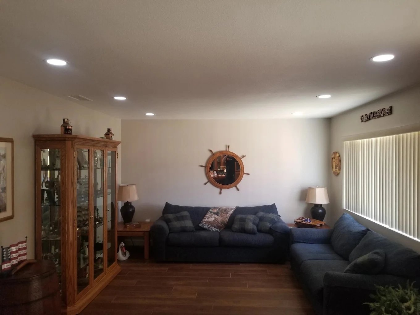 Living room with evenly spaced recessed ceiling lights above blue sofas, a wooden display cabinet and a ship's wheel mirror