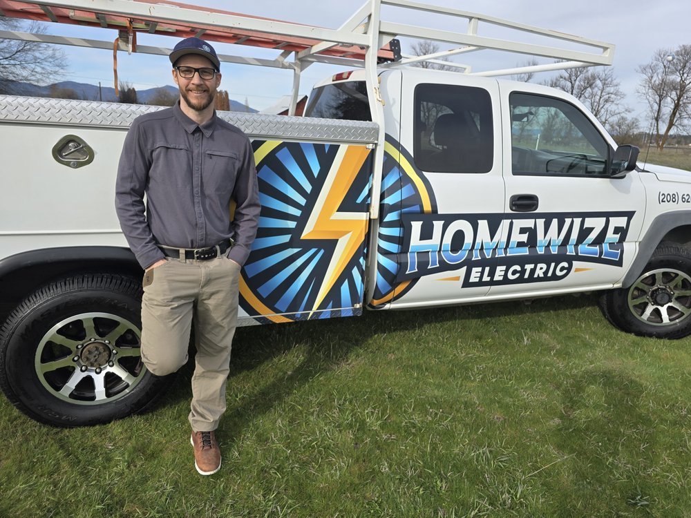 Smiling electrician in cap and glasses leans against a white service truck with Homewize Electric lightning-bolt logo and ladder rack