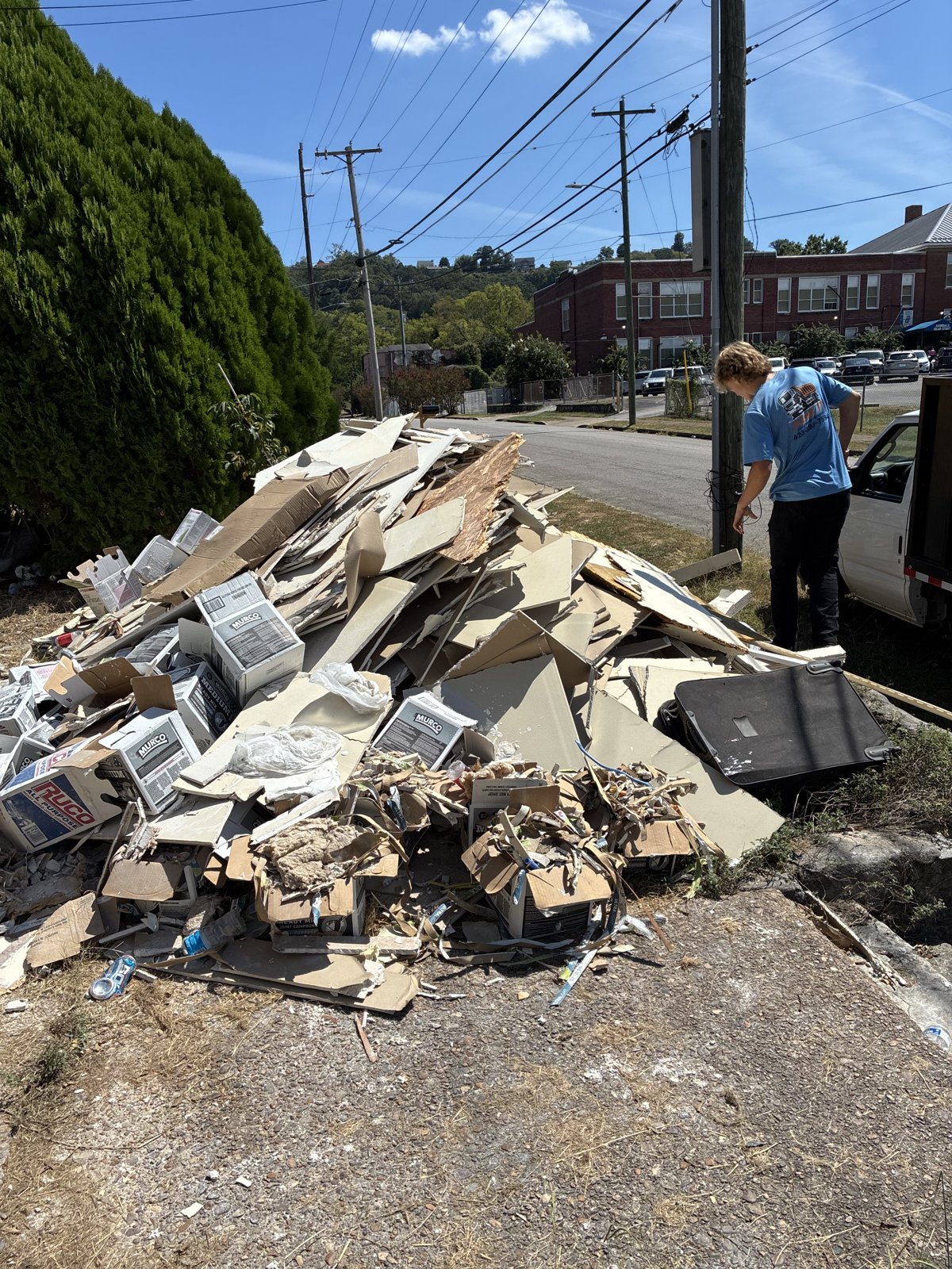 A Junk It worker in a blue shirt stands beside a large roadside pile of drywall scraps, cardboard boxes and other debris awaiting pickup.