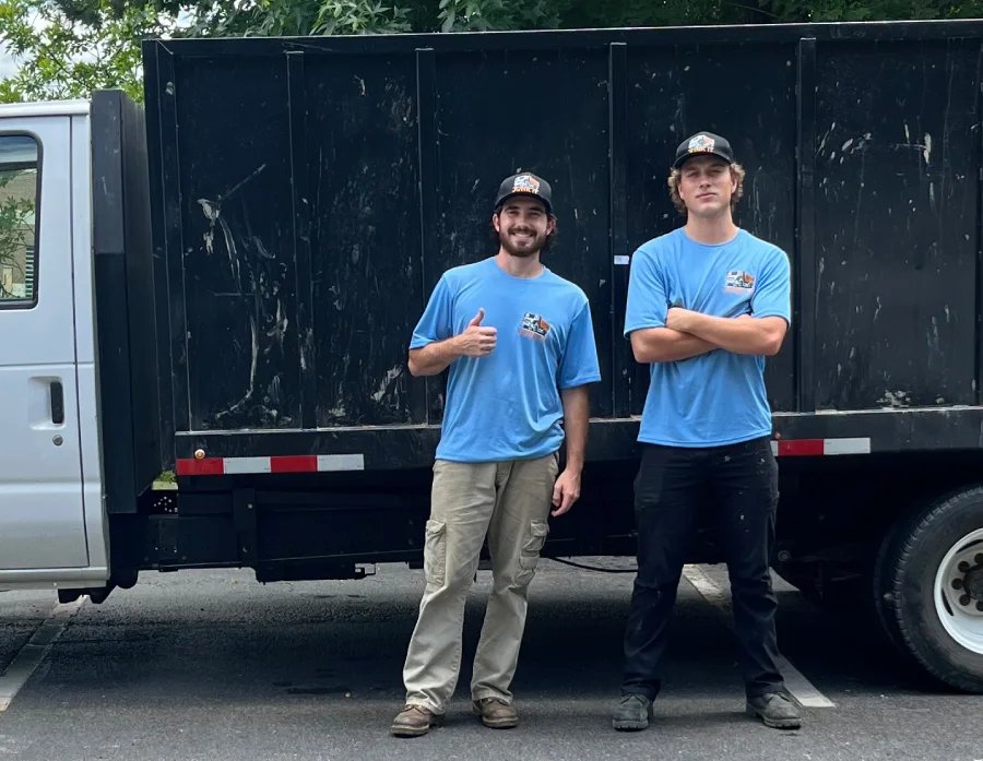 Two crew members in matching blue Junk It shirts stand in front of a black flatbed junk removal truck, one giving a thumbs up.