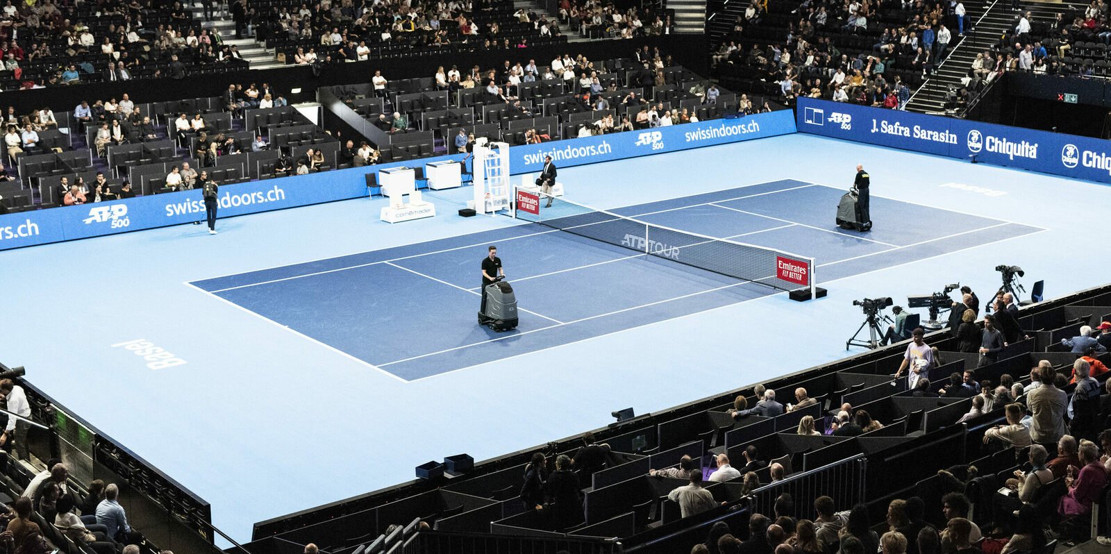 Operators with floor-cleaning machines work on a blue indoor tennis court as spectators fill the arena stands