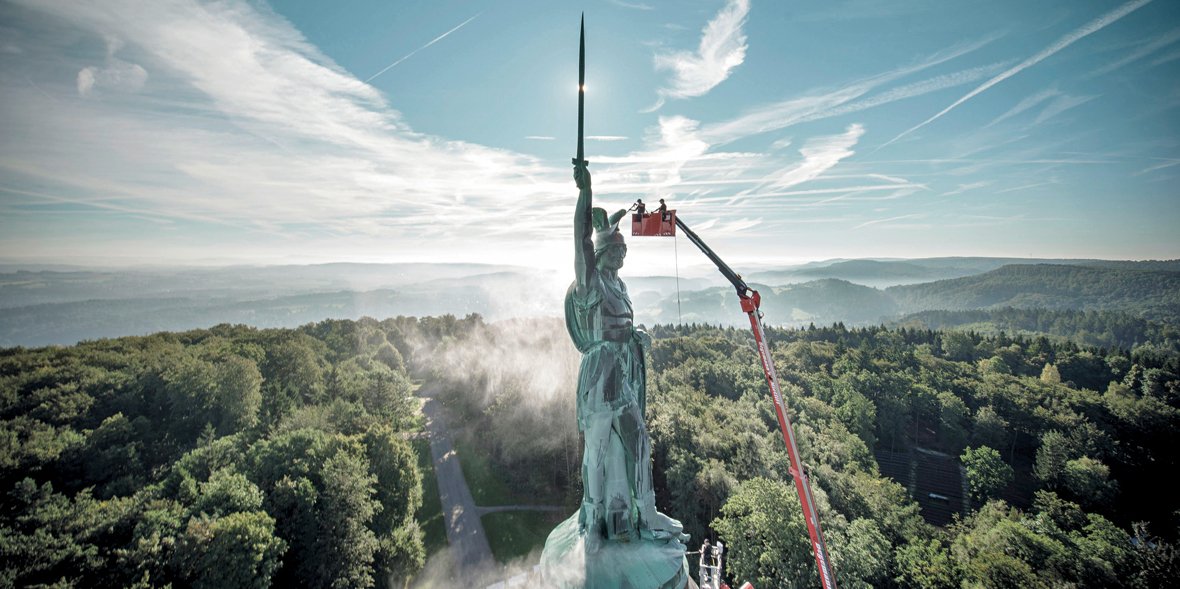 Workers in a red lift basket clean a large green bronze statue towering above a forested hilltop