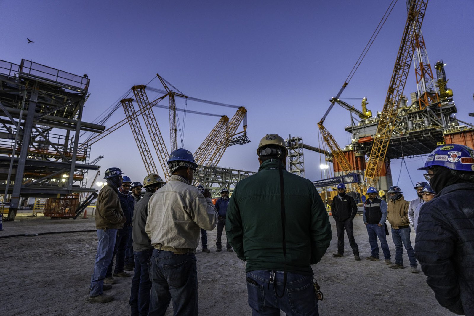 Crew members in hard hats gather at dusk on a worksite beneath large cranes and offshore platform structures