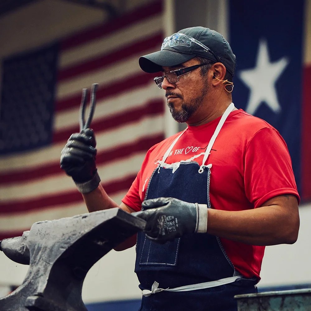 A worker in a red shirt and denim apron holds up a pair of pliers beside an anvil, with American and Texas flags hanging behind him