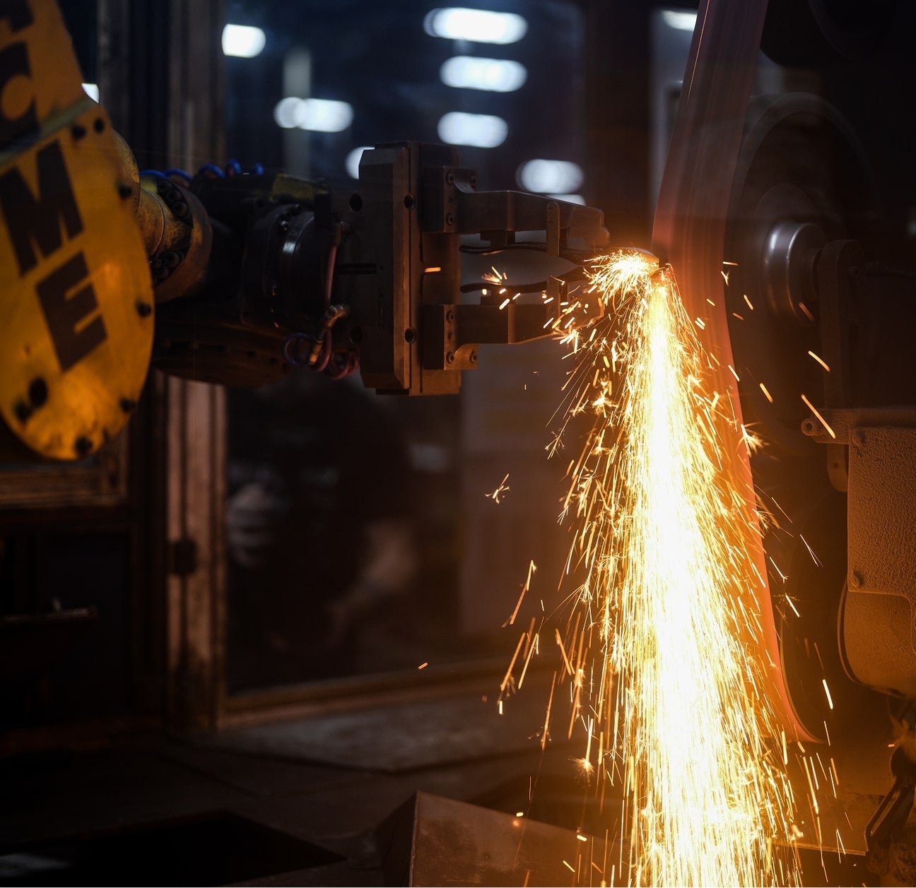 A yellow robotic arm holds a steel part against a spinning grinding belt, throwing a spray of sparks