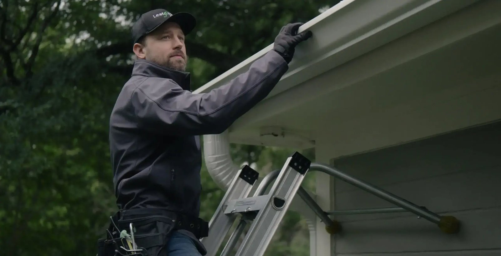 Installer in a dark jacket and LeafFilter cap stands on a ladder, reaching up to a home's gutter