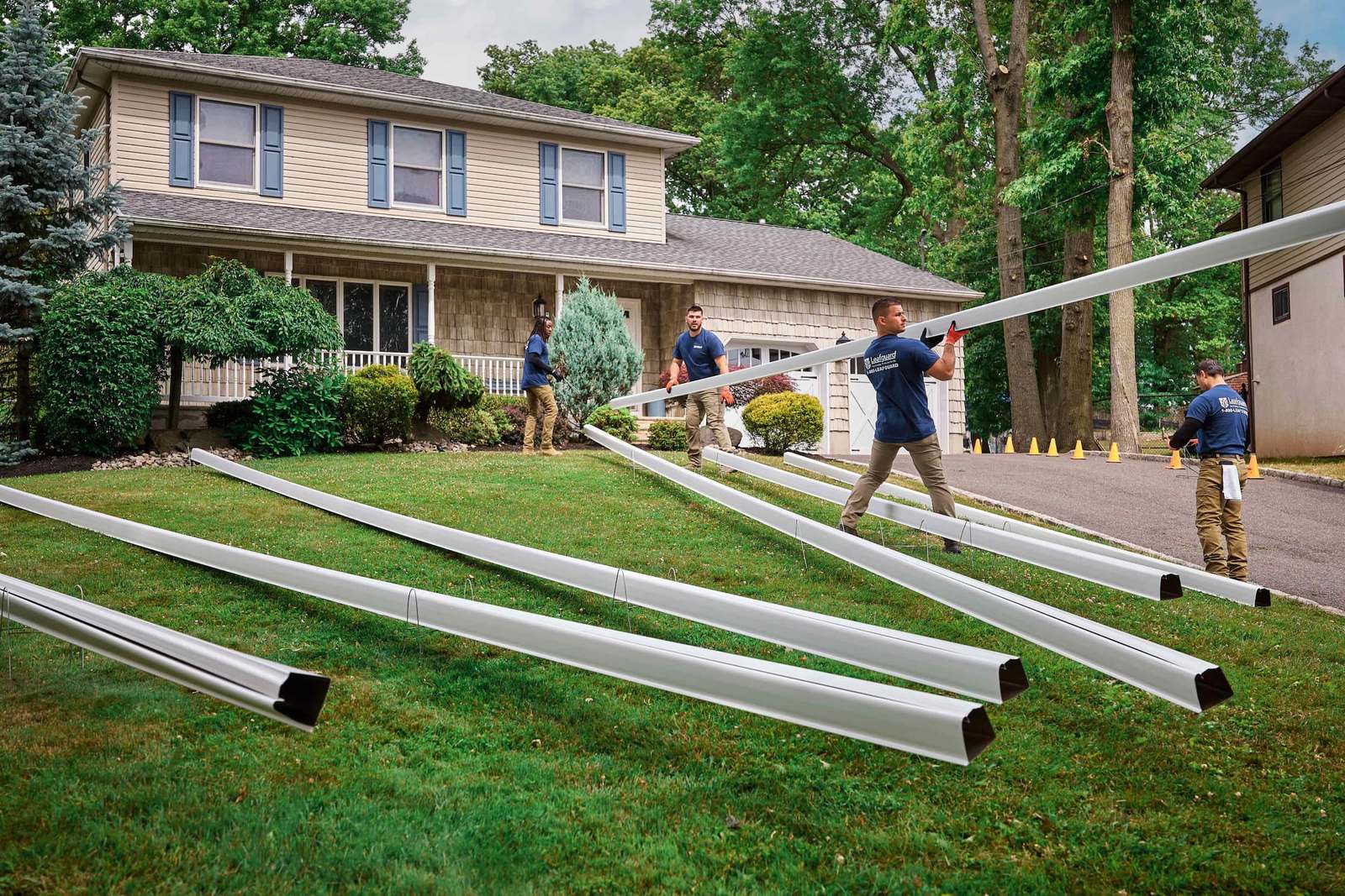 Four crew members in blue shirts carry long white gutter sections across a front lawn toward a house