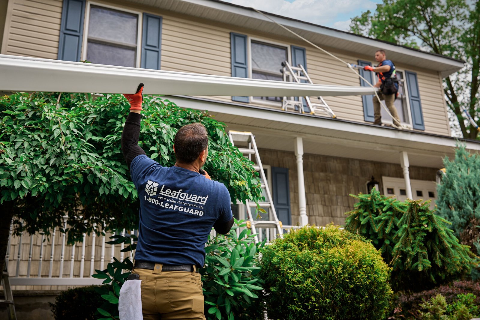 Installer in a Leafguard shirt lifts a long gutter section toward a second worker on a ladder at a two-story house