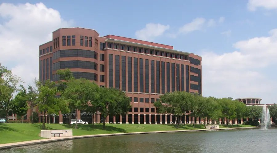Multistory red-brick office building with dark windows beside a landscaped pond with a fountain on a sunny day