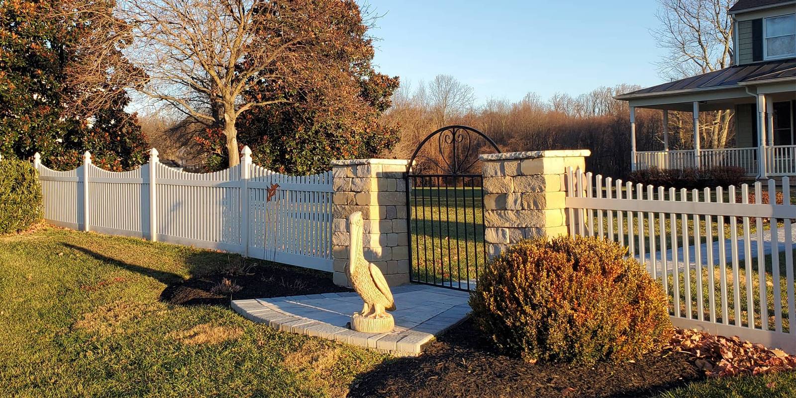 White vinyl picket fence and stone pillars with an arched black iron gate beside a garden pelican statue