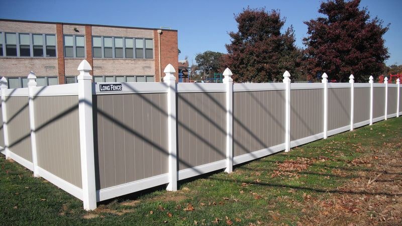 Tan and white vinyl privacy fence with capped posts running along a lawn beside a brick school building
