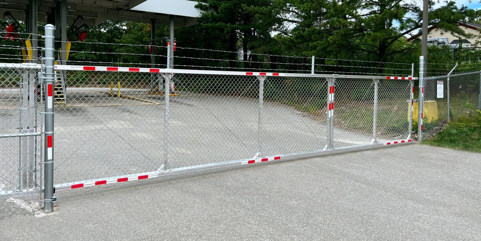 Galvanized chain link gate with red and white striped markers beside a paved lot, topped with barbed wire